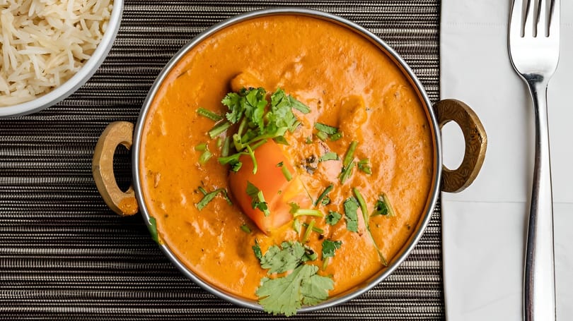 Overhead view of a vibrant orange curry in a metal bowl, garnished with cilantro and a tomato wedge.