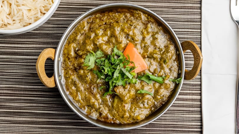 A metal bowl of green curry with cilantro and a carrot slice, served with white rice.